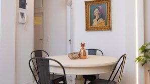 Spiseområde med et rundt bord, fire stole, et billede på væggen og en plante til siden.