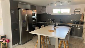 A modern kitchen with a stainless steel refrigerator, built-in oven, microwave, and a dining area with a white table and wooden stools. - Peace and comfort (Cessenon-sur-Orb)