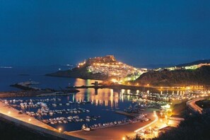 Una cittadina costiera di notte, con un castello in posizione elevata, un porto turistico con barche e un lungomare illuminato.