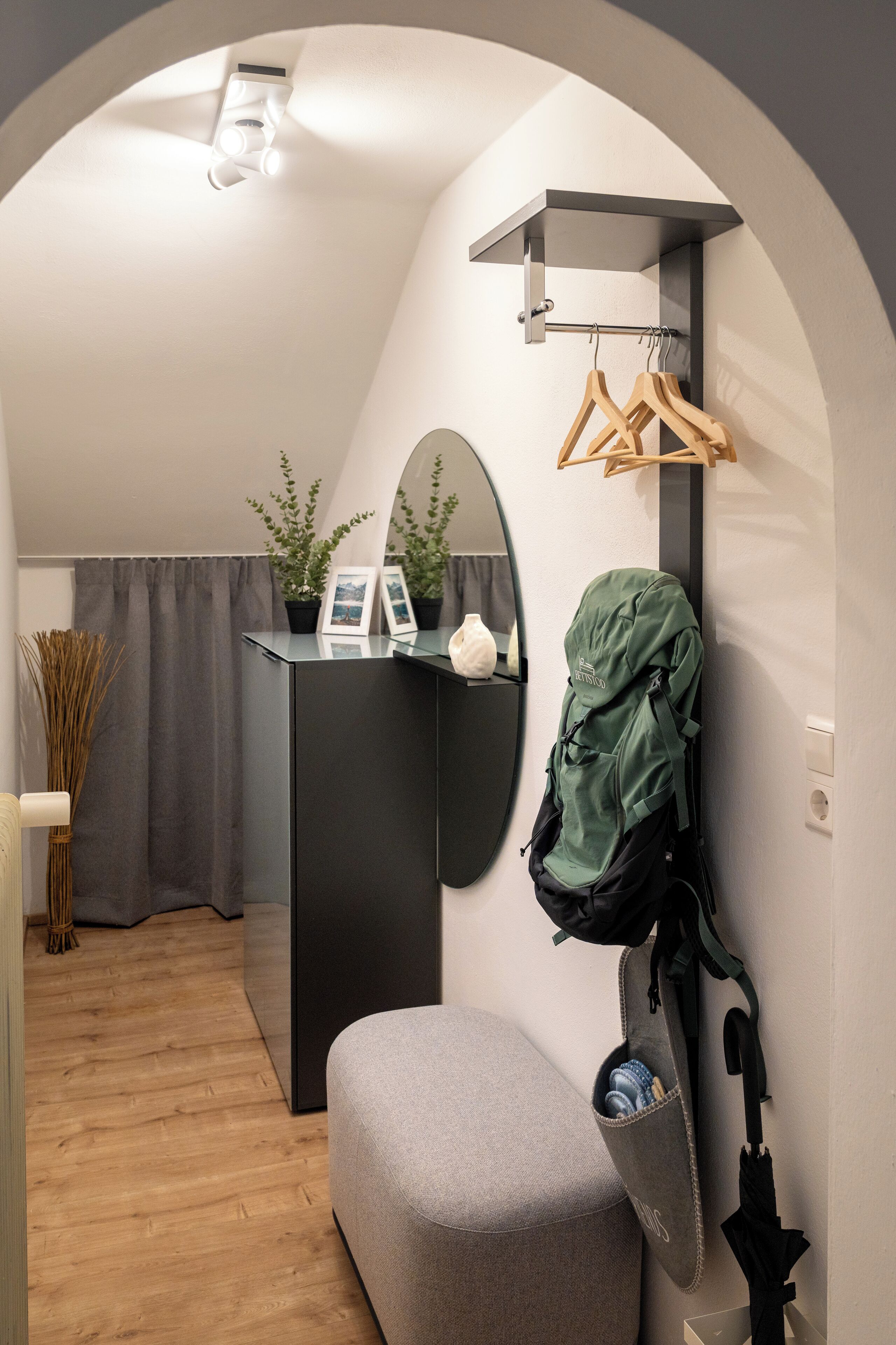 Una habitación pequeña con un banco, un espejo, una mochila verde y un paraguas colgando.