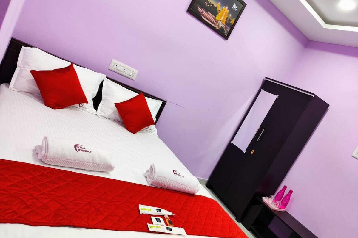 A hotel room with a bed, pillows, a red blanket, a minibar, and a mirror.