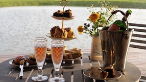 A table set with a variety of pastries, a bottle of champagne, and a flower vase by a lakeside. - Wilderness Reserve - Poachers (Halesworth)