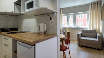 Cocina compacta con encimera de madera, microondas, hervidor y lavavajillas. Hay una zona de comedor con una silla y un sofá. Se ve una ventana con cortinas, y se observa un edificio rojo en el exterior.