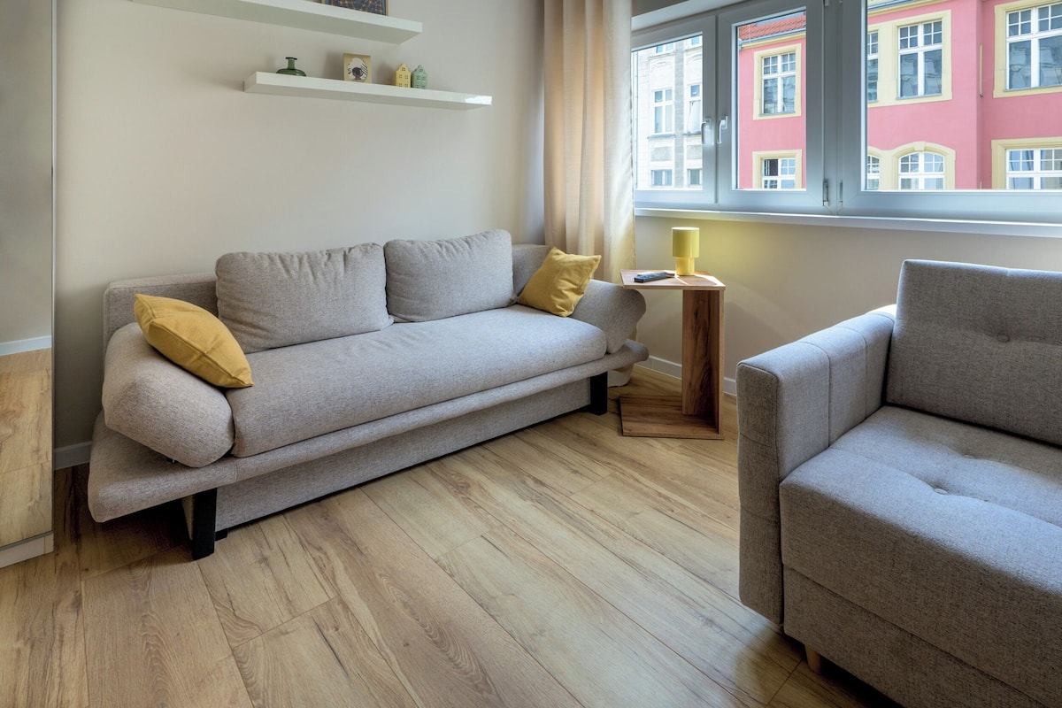 A modern living room with a grey sofa, a wooden side table with a lamp, and a view of a pink building through a window.