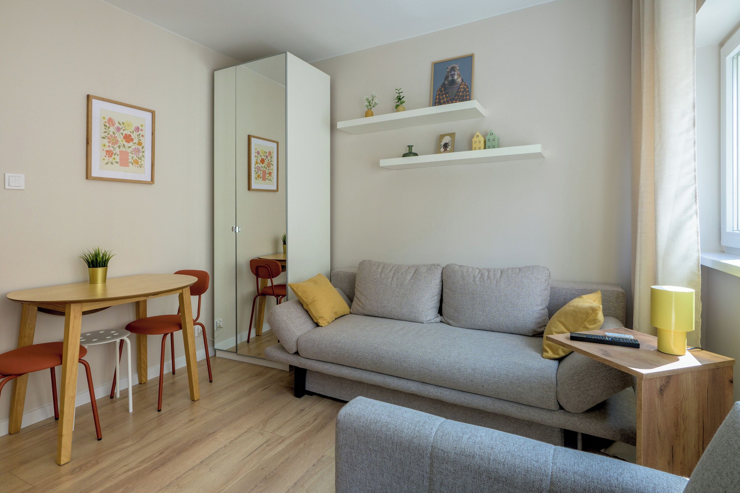 Uma sala de estar moderna com um sofá cinzento, uma mesa de centro de madeira e cadeiras vermelhas. Há estantes brancas com objetos decorativos e um espelho na parede.