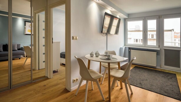 Una sala de estar moderna con comedor, televisor de pantalla plana en la pared y ventanas grandes que ofrecen luz natural.