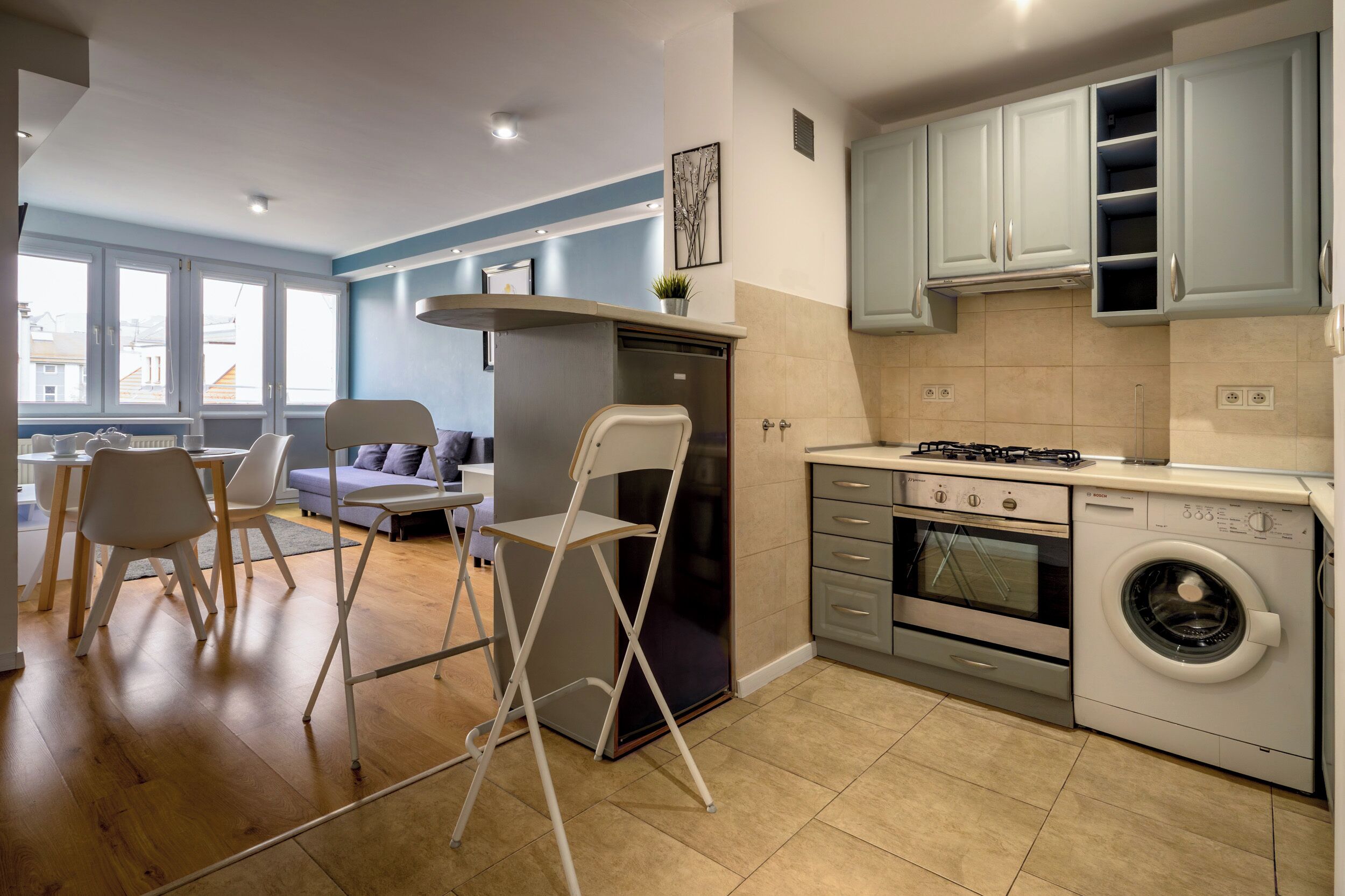 A modern kitchen with a gas stove, oven, and washing machine. There is a dining area with chairs and a table.