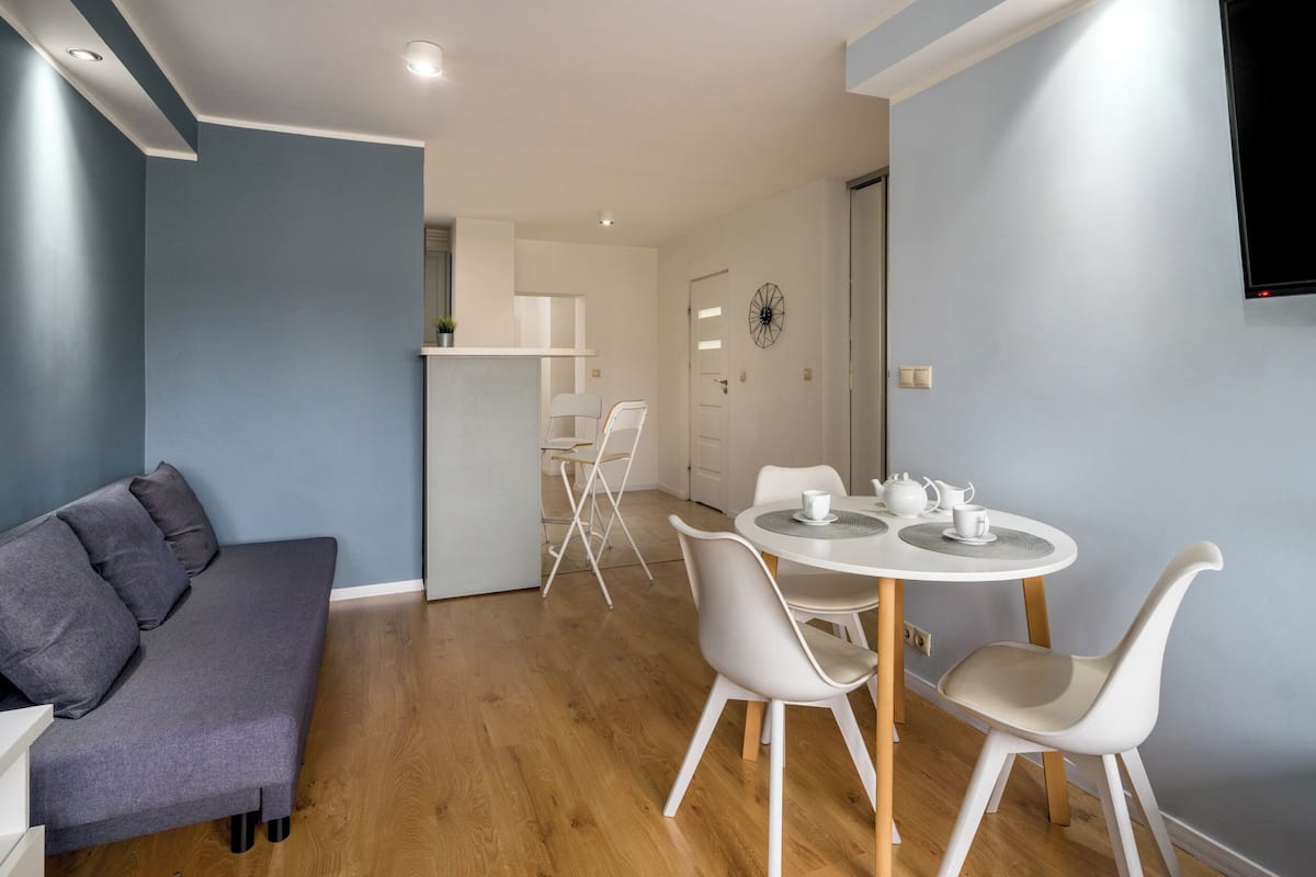 A modern living room with a grey sofa, a round dining table with white chairs, and a kitchen area in the background.