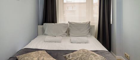 Una cama bien hecha con sábanas blancas y dos almohadones decorativos, una ventana con cortinas y una pared de color azul claro.