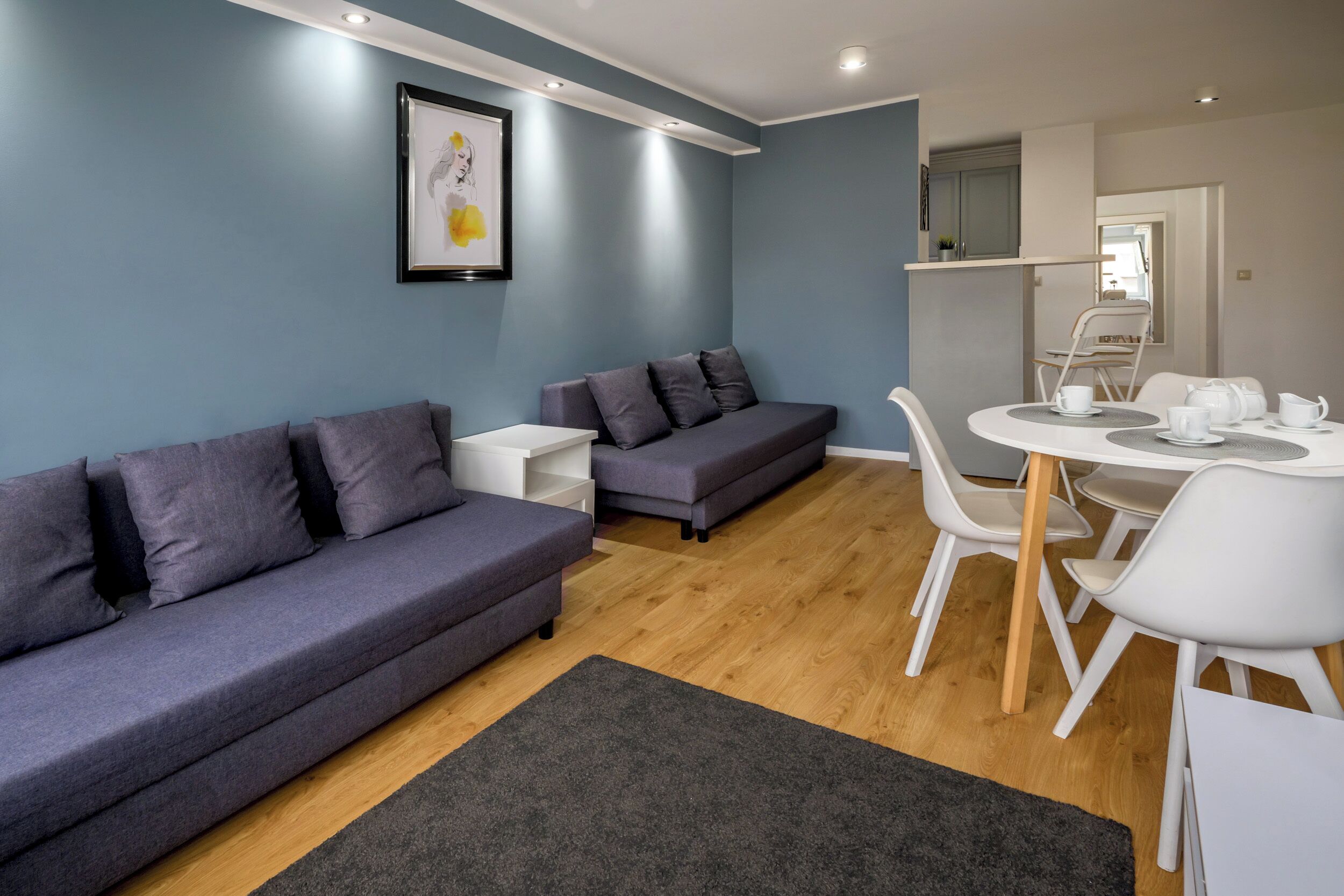 A modern living room with a grey sofa, a smaller grey sofa, a white table with chairs, and a framed picture on the wall.