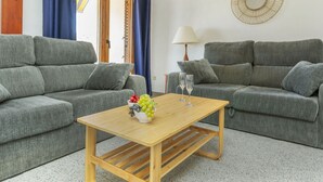 Una sala de estar con dos sofás grises, una mesa de centro de madera con un cuenco de fruta y dos copas de vino, y una lámpara en una mesita auxiliar.