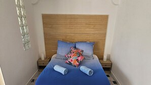A neatly made bed with a blue bedspread, two pillows, and two rolled towels. The room has a wooden headboard, a small bedside table, and a tiled floor with a black and white pattern. - Vue MER bleu sea beach Holidays tramway (Nice)