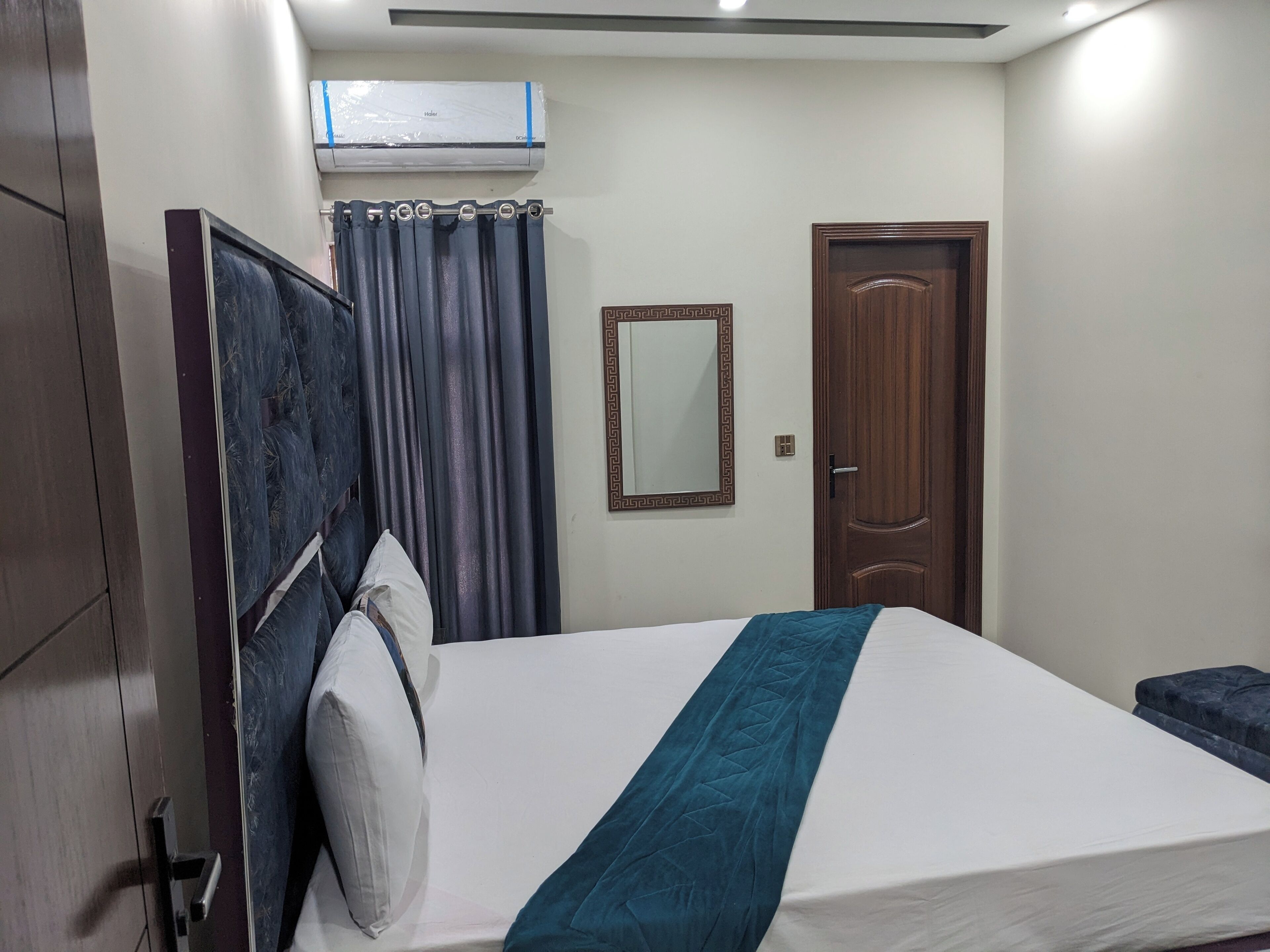 Una habitación de hotel con una cama, una puerta de madera, un espejo y un equipo de aire acondicionado.