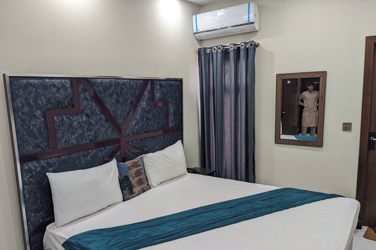 A neatly made bed with white and teal bedding, a headboard with a geometric pattern, a grey curtain, and a wall-mounted air conditioner.
