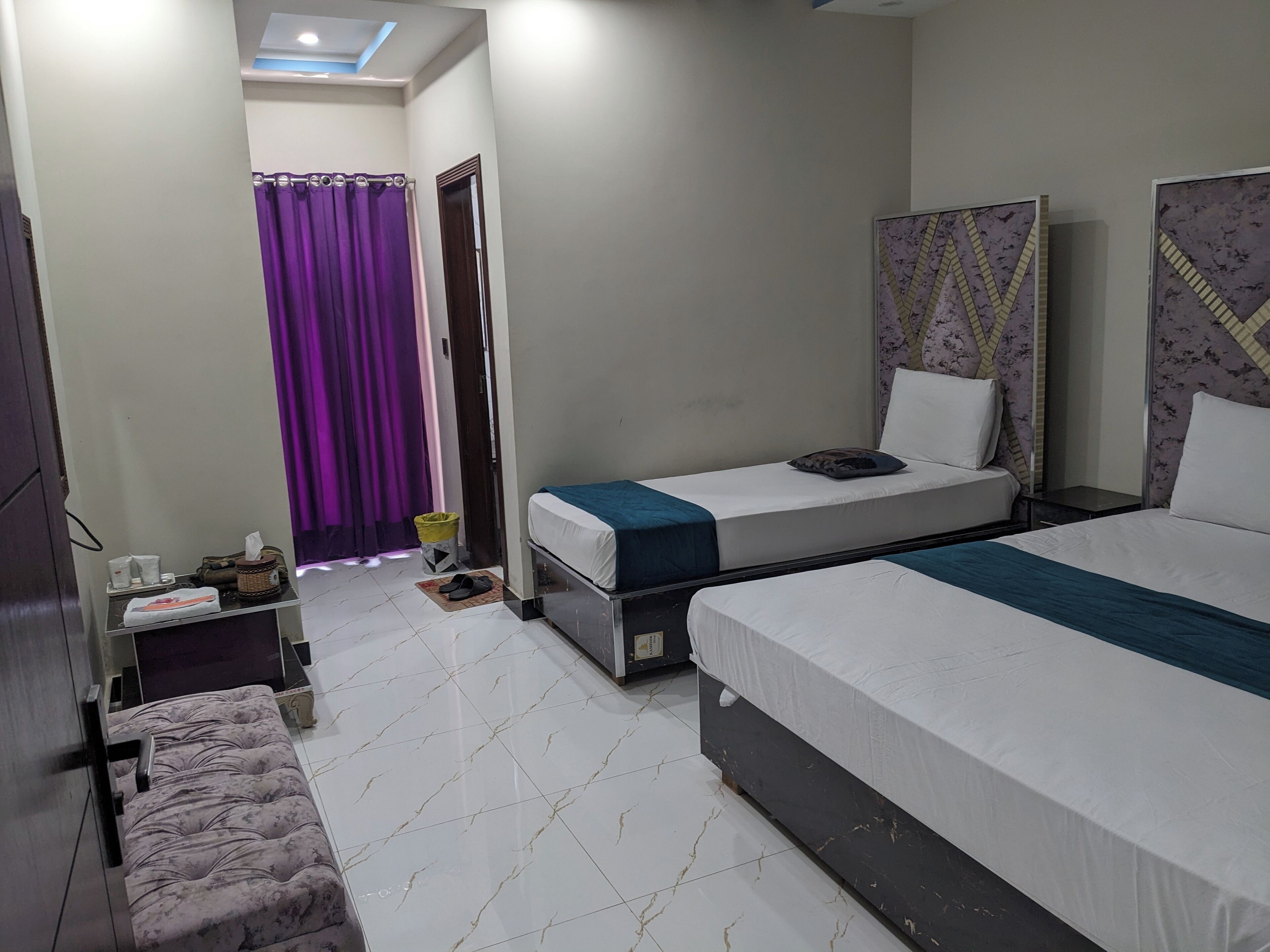 A hotel room with two beds, a small table with toiletries, a purple curtain, and a patterned headboard.