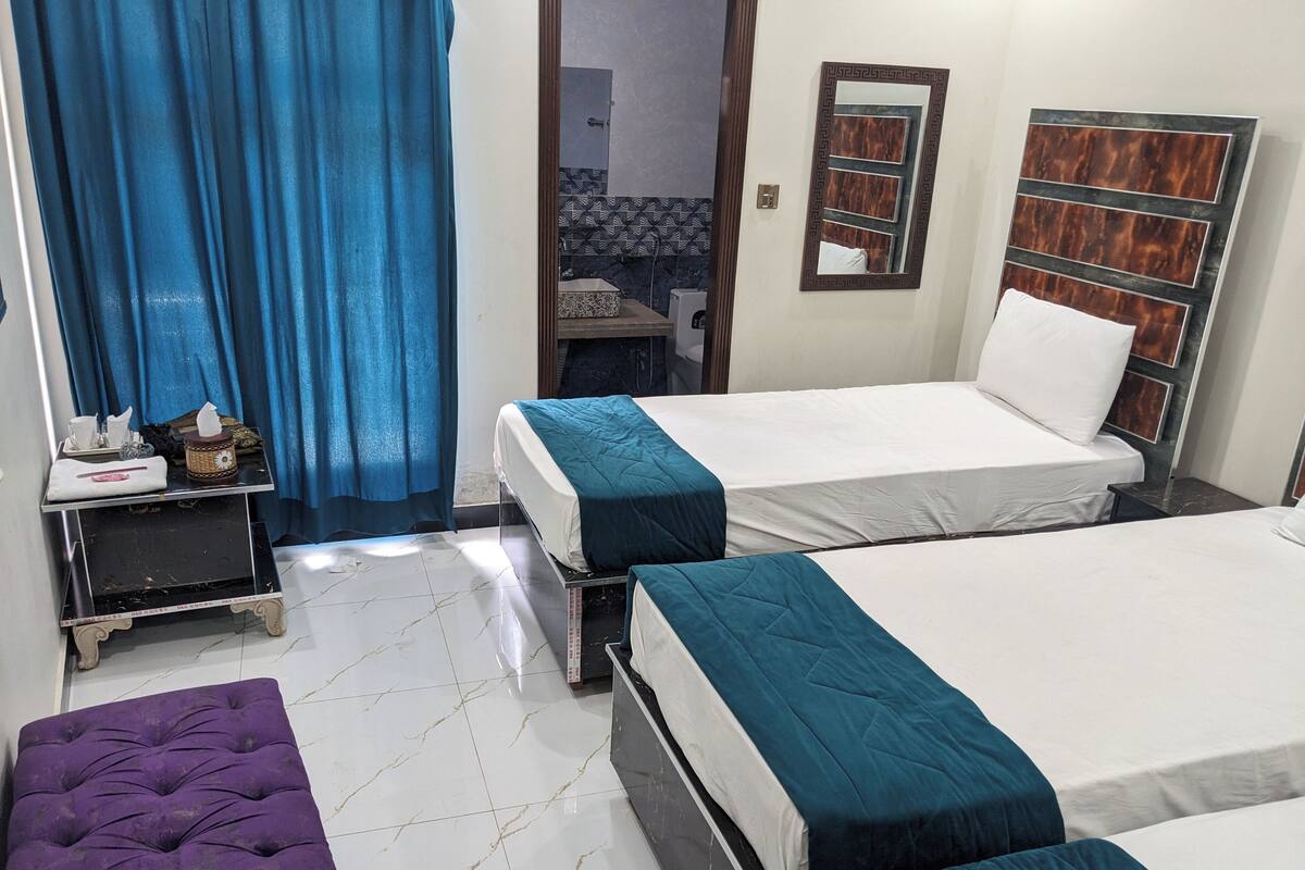 A hotel room with two beds, a small table with toiletries, a purple bench, and a bathroom visible through an open door.