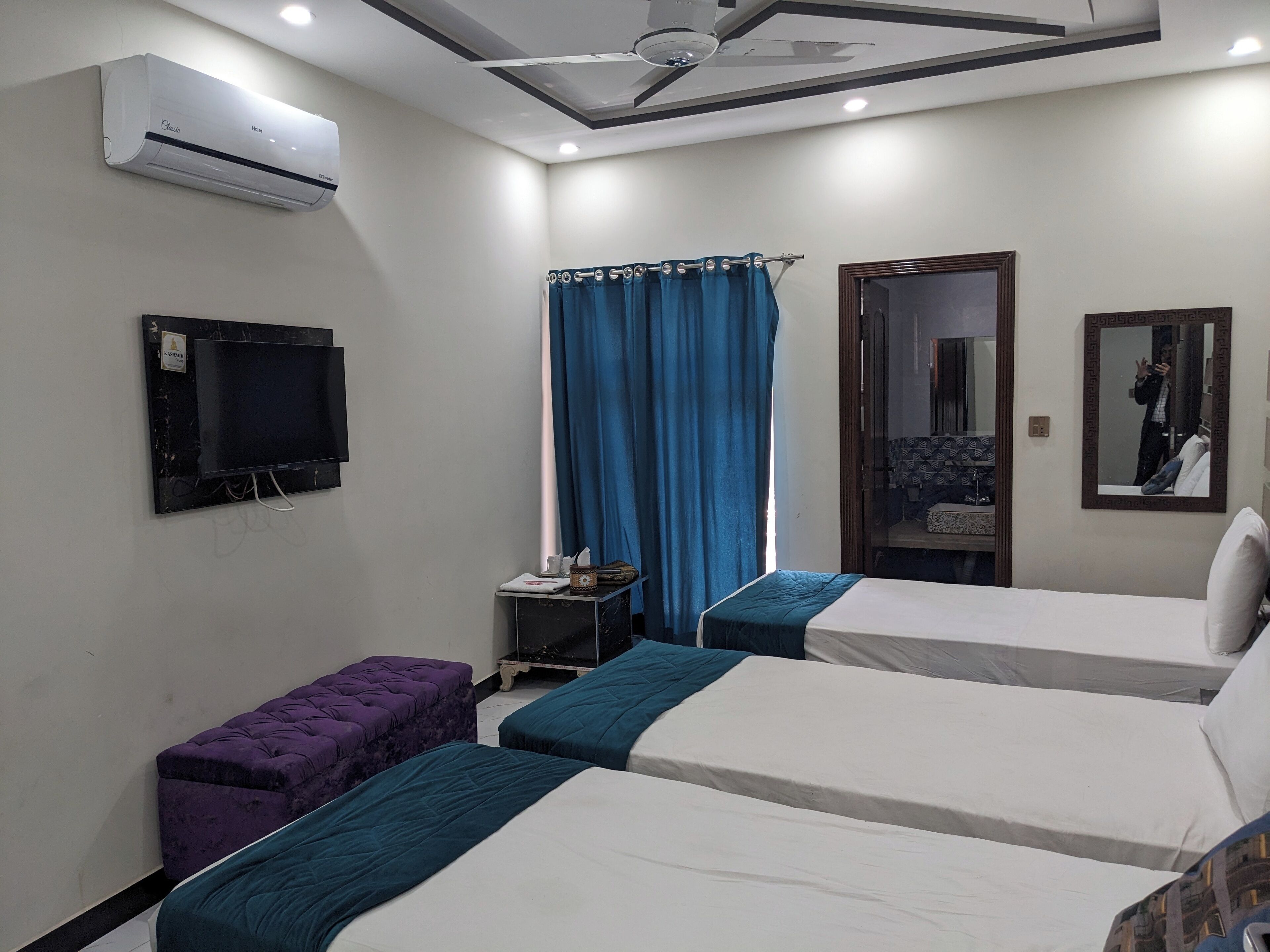 A hotel room with two beds, a TV, an air conditioner, a window with blue curtains, a small table with a tissue box, and a mirror on the wall.