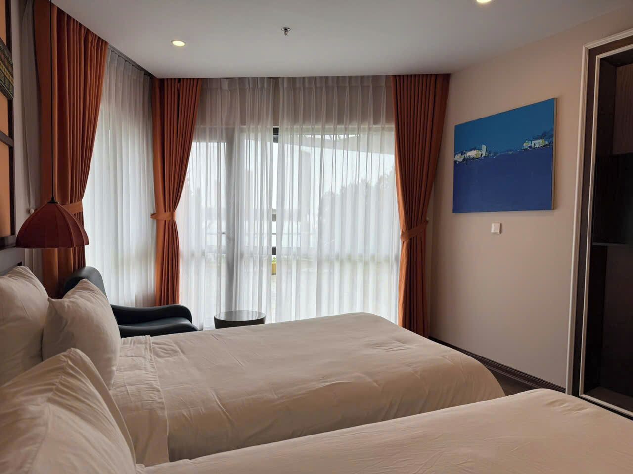 A hotel room with two beds, a desk, and a view of the outdoors through sheer curtains.