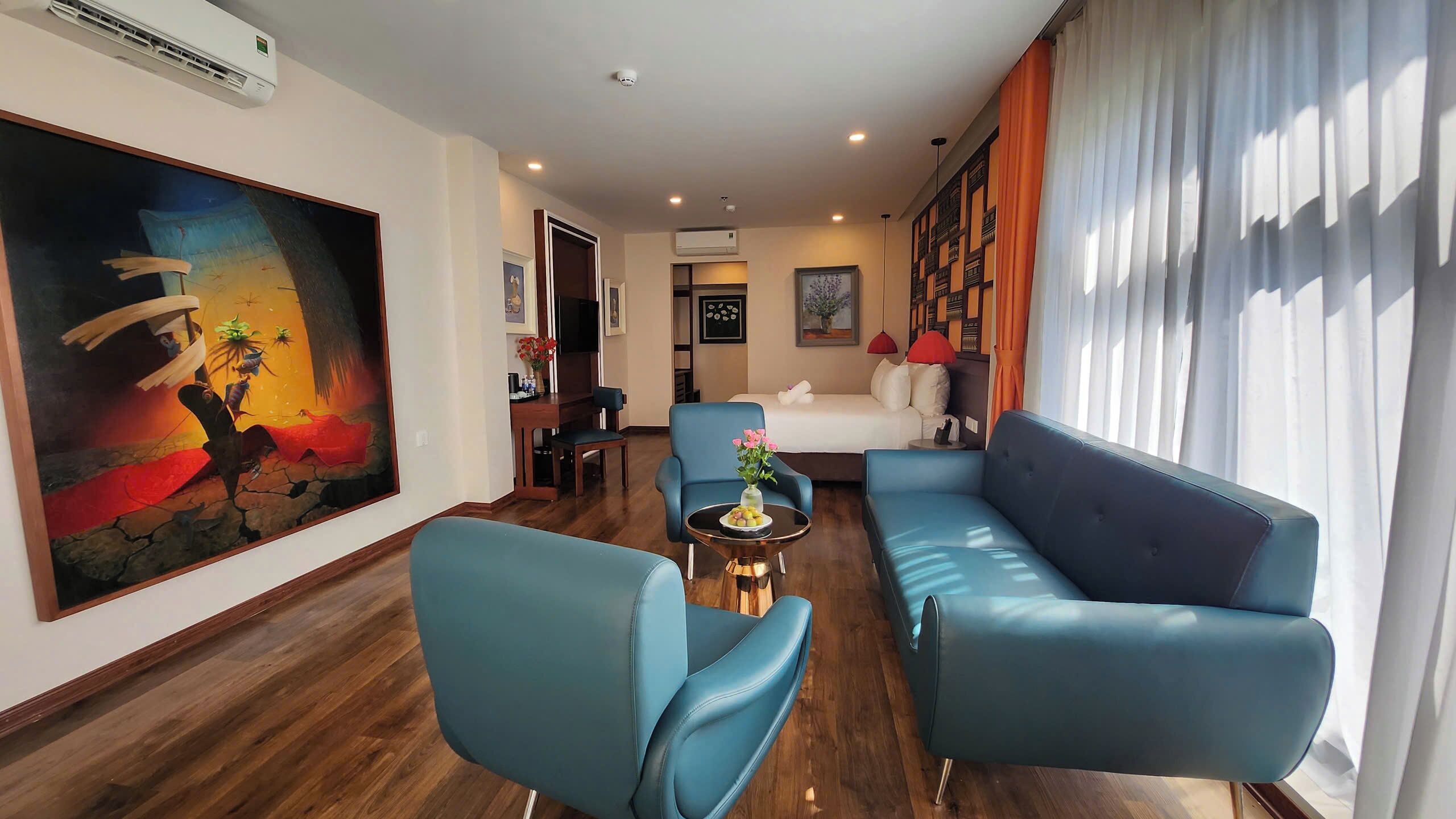 Una habitación de hotel moderna con un cuadro grande, un sofá, una silla, una mesita con flores y una cama con cabecera blanca.