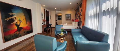 A modern hotel room with a large painting, a sofa, a chair, a small table with flowers, and a bed with a white headboard.