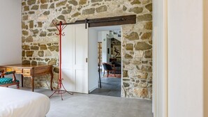 Una habitación con pared de piedra, una cama, un escritorio, una silla y un perchero.