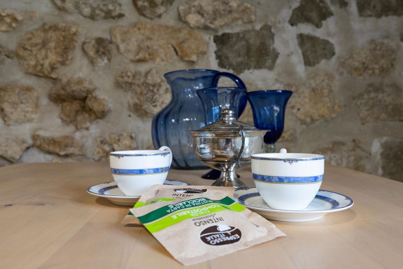 Une table en bois, adossée à un mur de pierre, sur laquelle on trouve deux tasses blanches ornées de motifs bleus, un pichet bleu, un récipient en argent et un paquet de café.