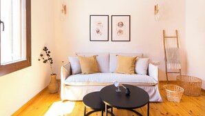 A cozy room with a sofa, two framed artworks, a small table, a vase with branches, and a window. - Old Town Charm-Cozy 2BR with Rooftop (Rethymno)