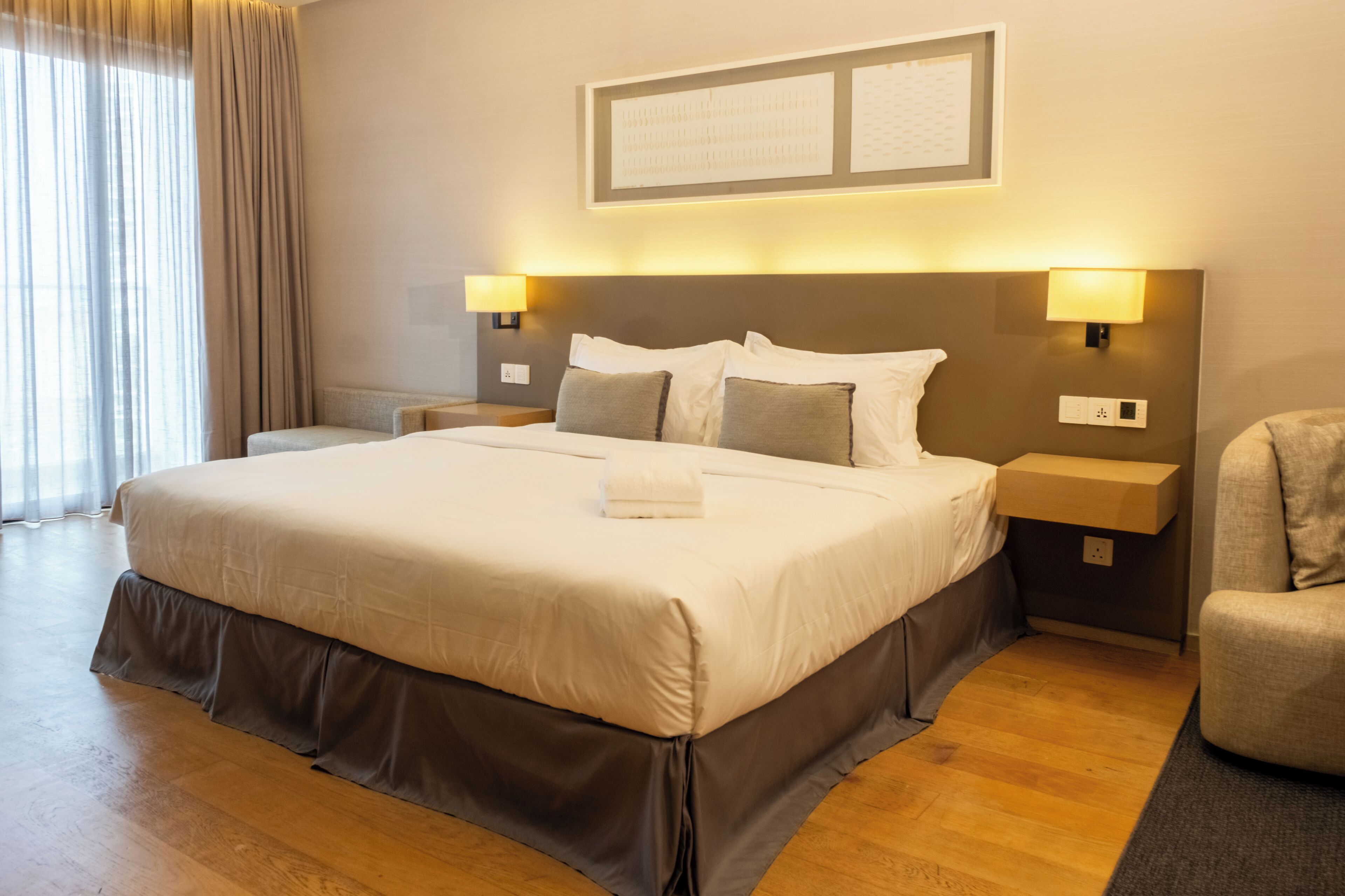 Una habitación de hotel con una cama grande, dos mesitas de noche, una silla y un ventanal con cortinas.