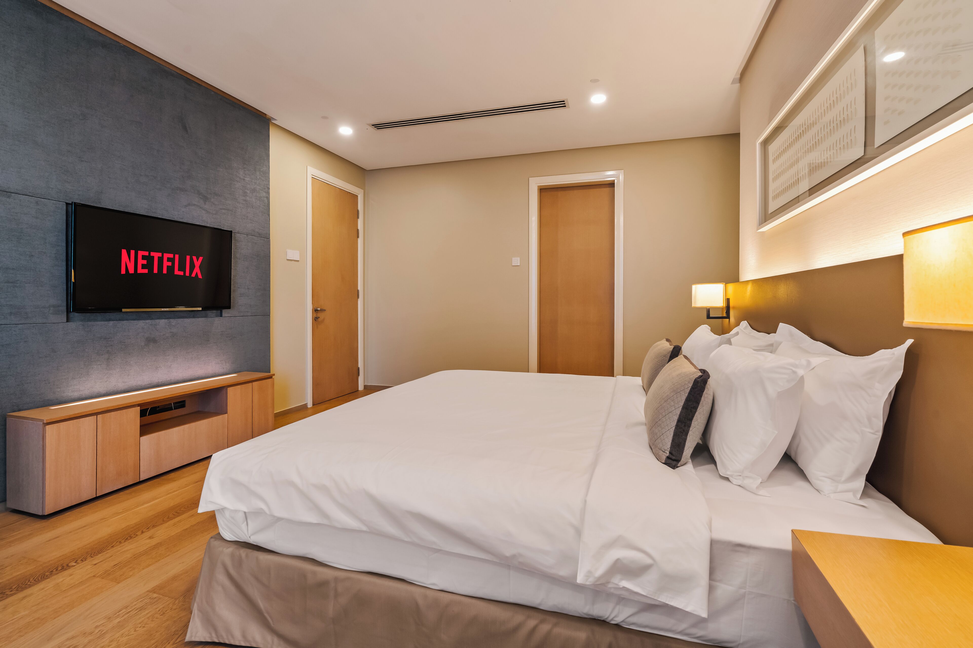 A modern hotel room with a large bed, a flat-screen TV mounted on the wall, a wooden bedside table, and a wall-mounted lamp.