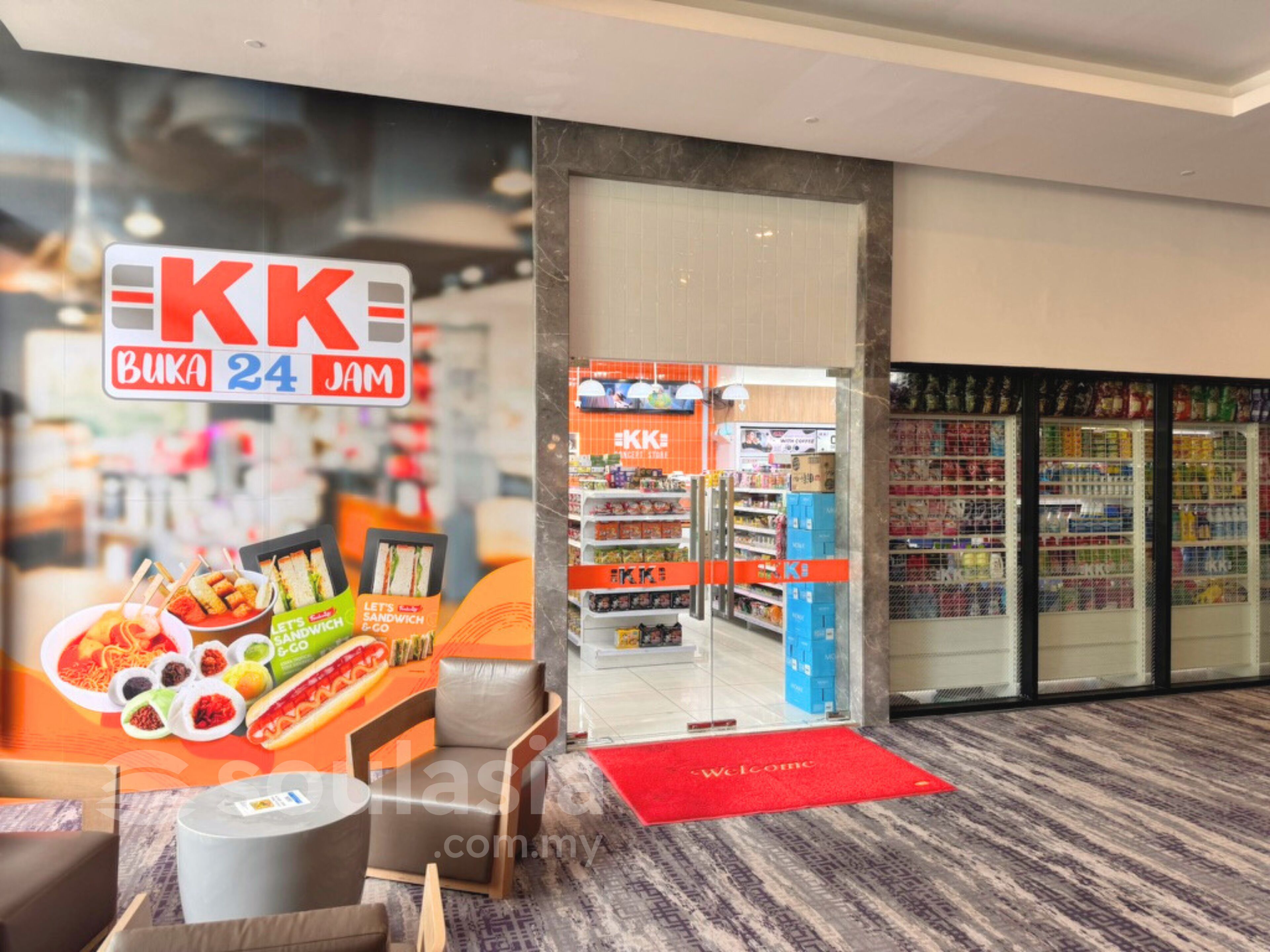 En butiksinredning med en skylt som visar texten ”KK BUKA 24 JAM” och ett utbud av livsmedel.