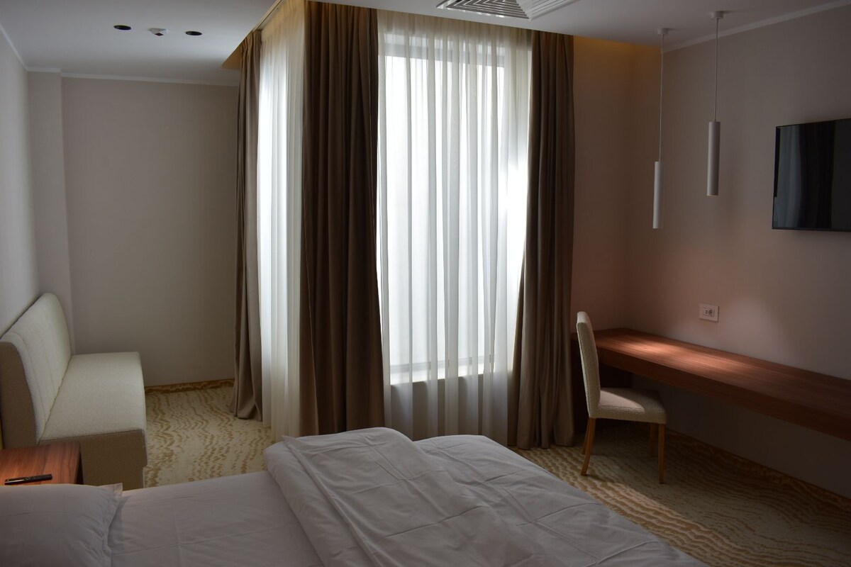 A hotel room with a bed, a desk, a chair, and a large window with curtains.
