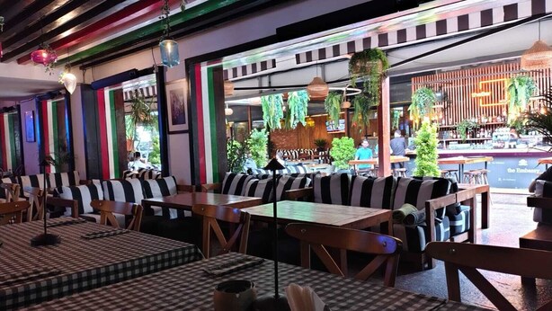 Un restaurante con manteles a cuadros, asientos acolchados a rayas, plantas colgantes y una zona de bar al fondo.