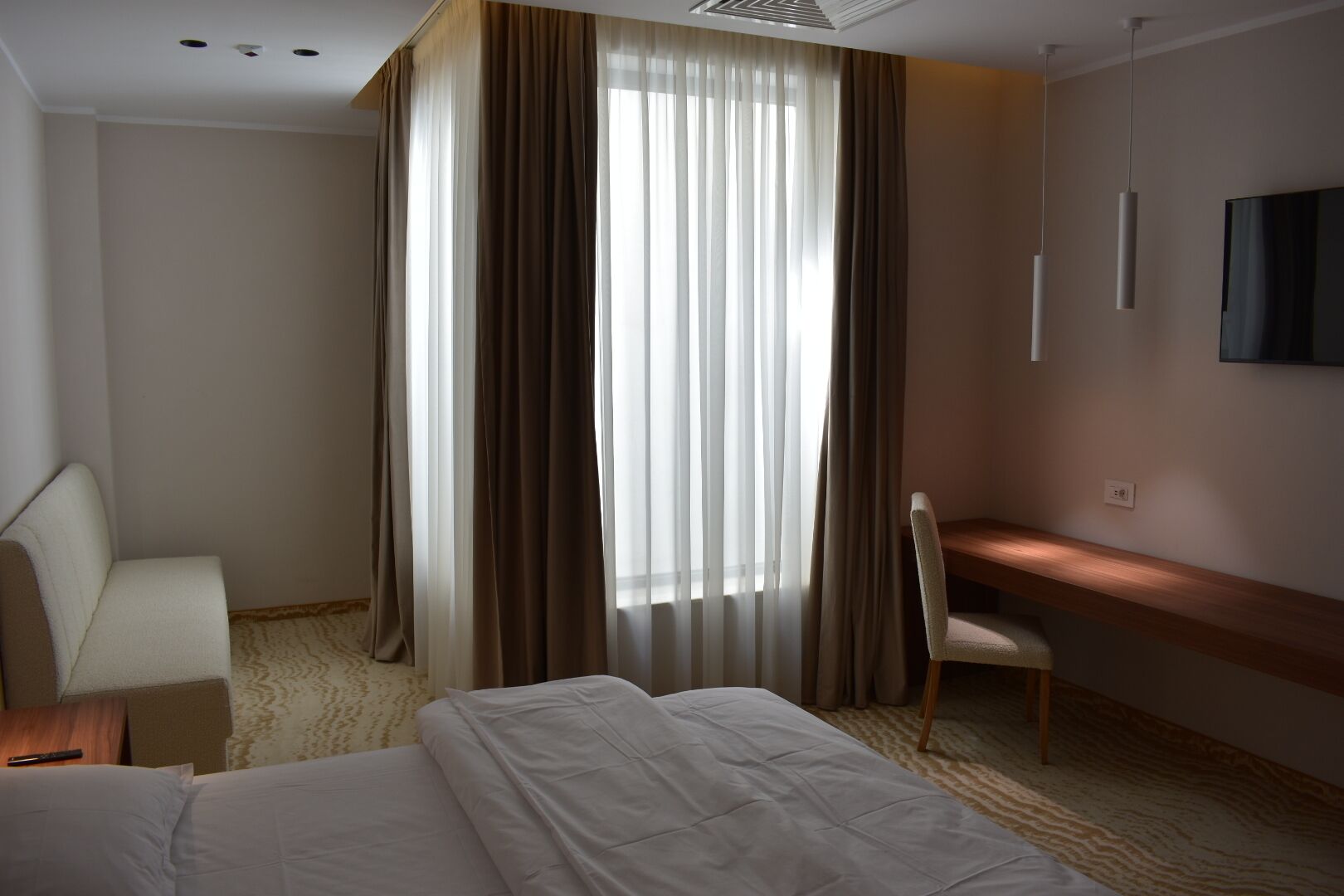 Una habitación de hotel con una cama, un escritorio, una silla y un amplio ventanal con cortinas.
