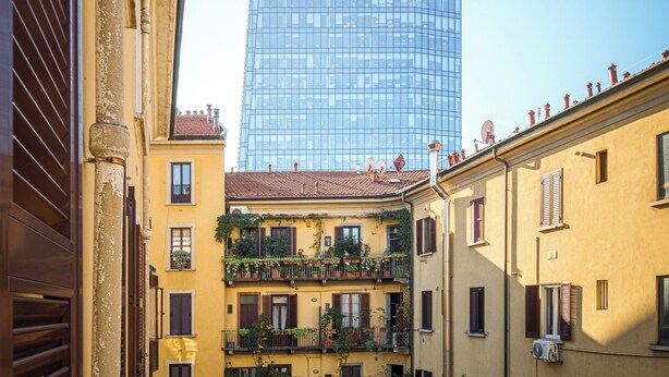 Vista de um edifício alto de vidro com o texto 'BNP PARIBAS', rodeado por edifícios amarelos com varandas.