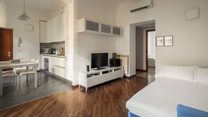 Quarto de hotel moderno com piso de madeira, cama, televisão, cozinha compacta e área de refeições.