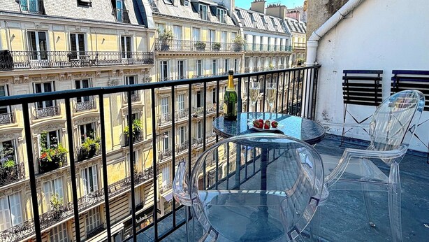 A rooftop terrace with a glass table and chairs, a bottle of champagne, and a view of Parisian buildings. - L'appartement Colombier (Paris)