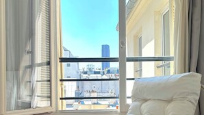 A modern room with a white cushioned chair, a large window with a view of buildings, and a wooden floor. - L'appartement Colombier (Paris)