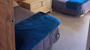 Een kamer met houten paneelwanden, twee bedden met blauw beddengoed, een klein houten dressoir en een raam met gordijn.