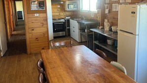 Een keuken met houten lambrisering, een eettafel, stoelen, een magnetron, fornuis, spoelbak en een koelkast.