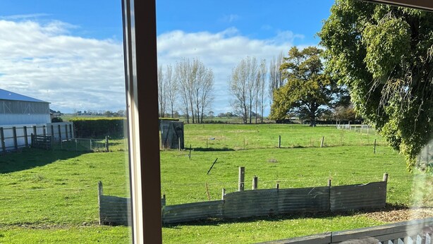 Uitzicht vanuit een raam op een grasveld met een houten omheining, een gebouw op de achtergrond en een heldere lucht.