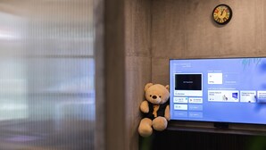 Ein Zimmer mit Fernseher, der eine Benutzeroberfläche für ein Smart Home zeigt, einem Teddybären und einer Uhr an der Wand.