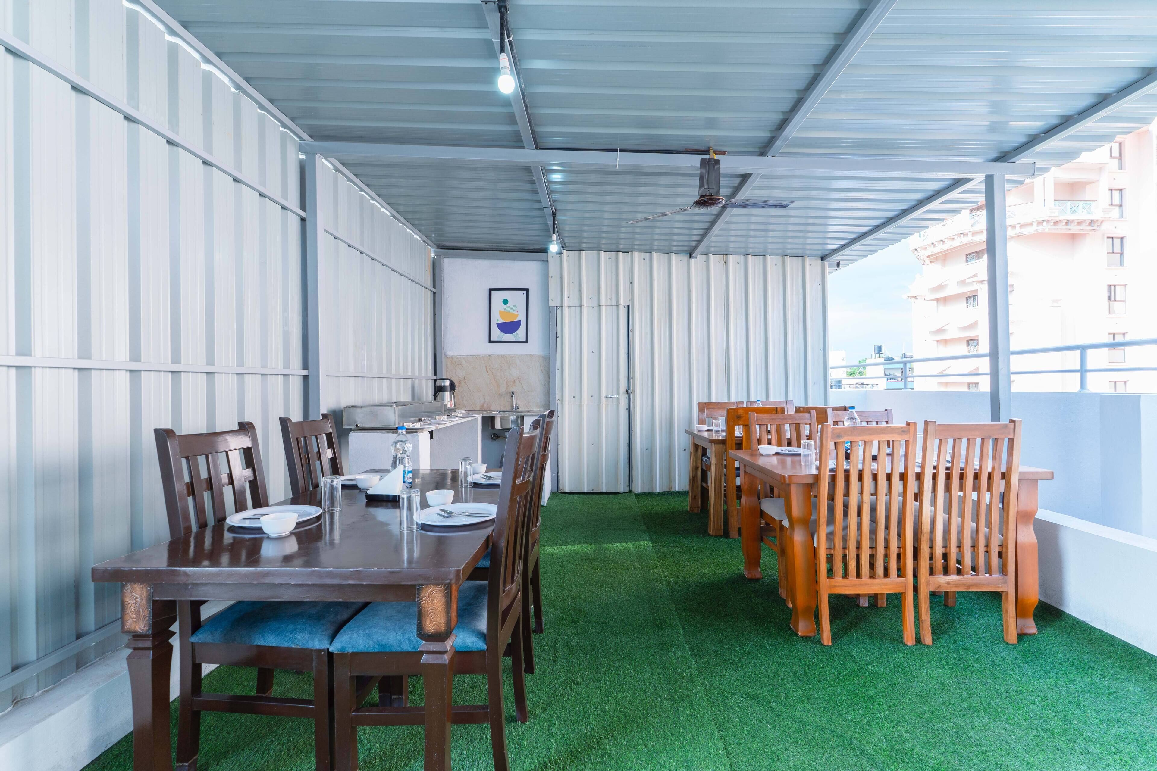 Un restaurante con un área exterior techada que cuenta con césped artificial verde, mesas y sillas de madera, y vistas de edificios al fondo.