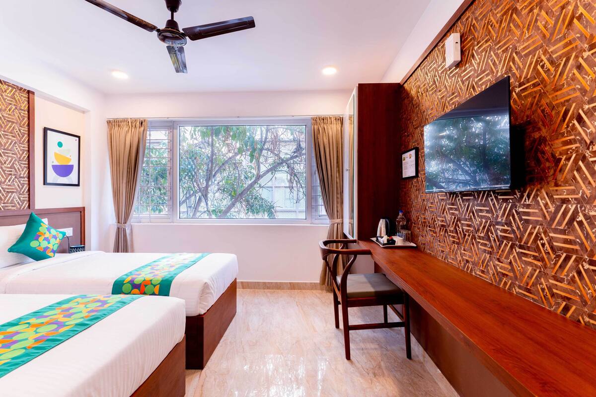 A hotel room with two beds, a desk, a television, and a window with a view of trees.