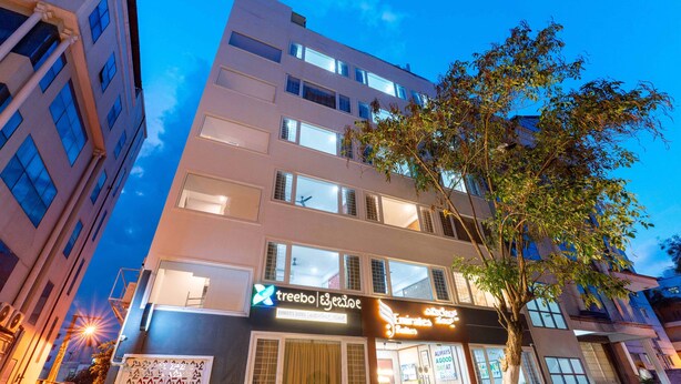 A multi-story hotel with a modern facade, large windows, and a visible tree in front. - Treebo Emirates Suites Indiranagar (Bangalore)