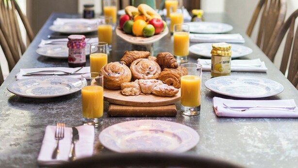 Une table de petit-déjeuner dressée avec des assiettes, des croissants, des fruits et des verres à jus.