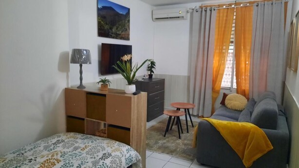 A small, well-lit room with a bed, a sofa, a desk with a lamp, a small table, and a window with curtains.