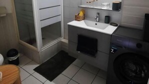 A compact bathroom with a white sink, a mirror, a towel rack, a washing machine, and a glass shower enclosure.