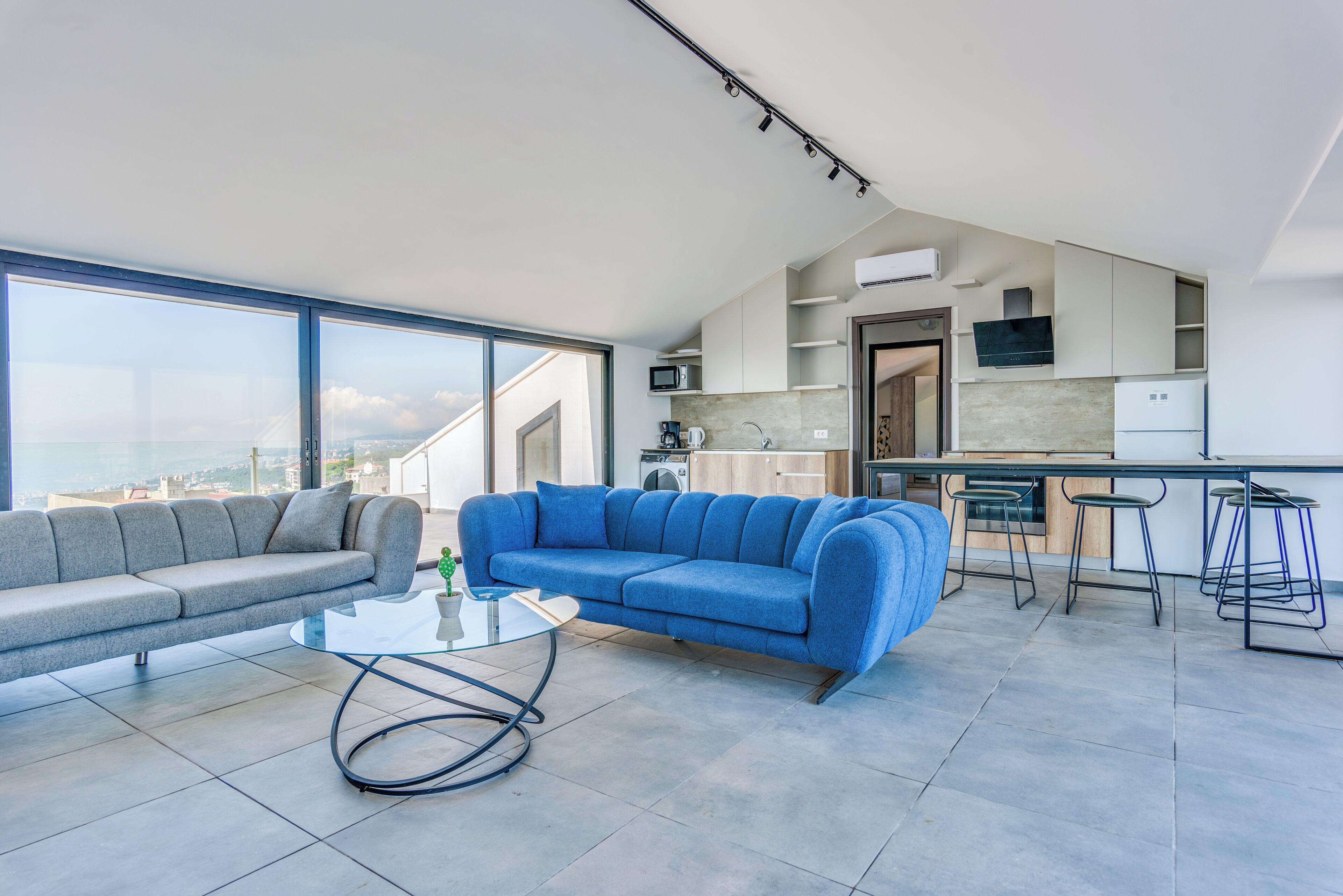 A modern living room with a gray sofa, a blue sofa, and a glass coffee table. There is a kitchen area with a microwave, stove, and bar stools. The room has large windows offering a view of the city.