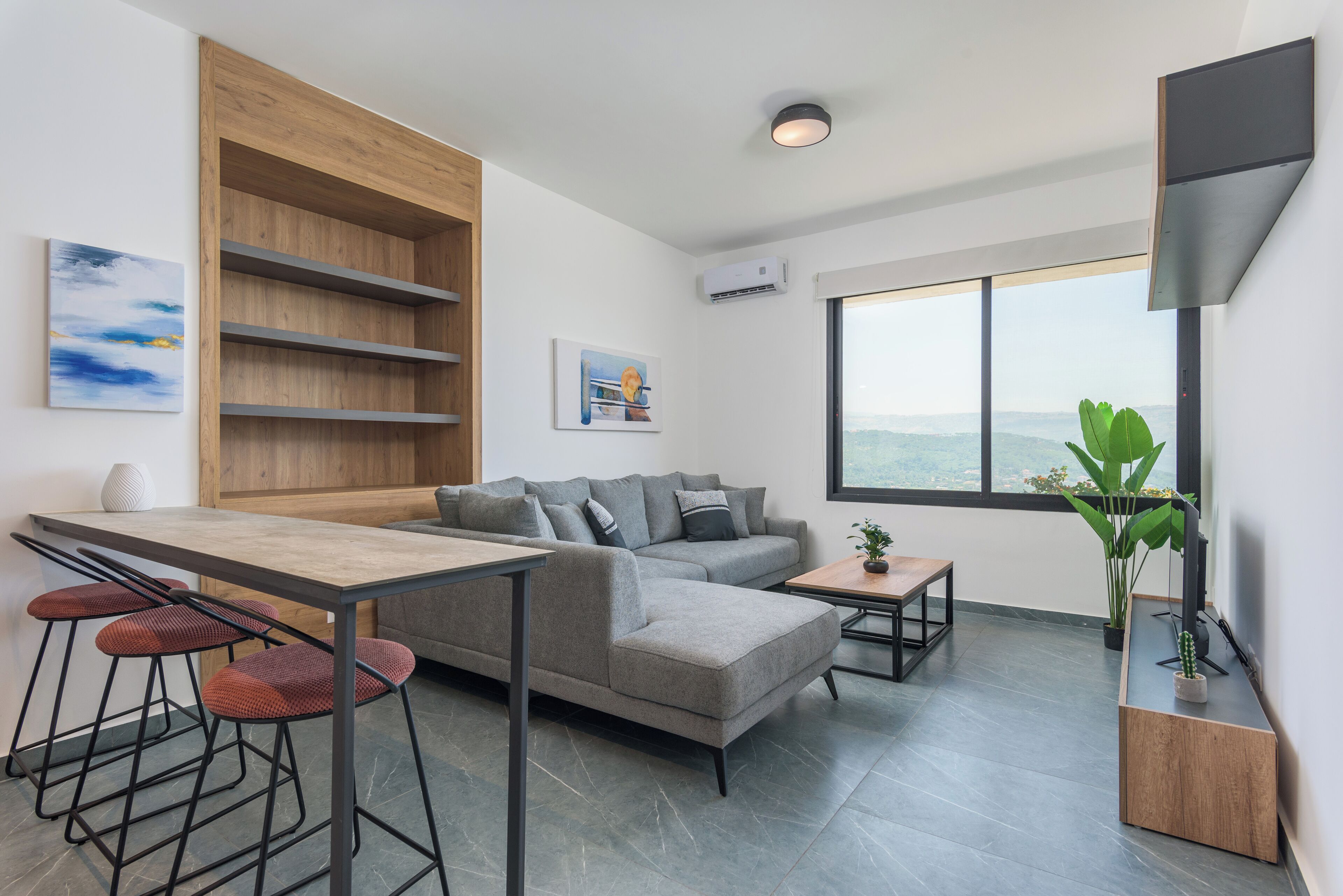 Um espaço de estar moderno com um sofá modular cinzento, uma estante embutida de madeira, uma mesa de jantar com banquetas altas e uma grande janela com vista.