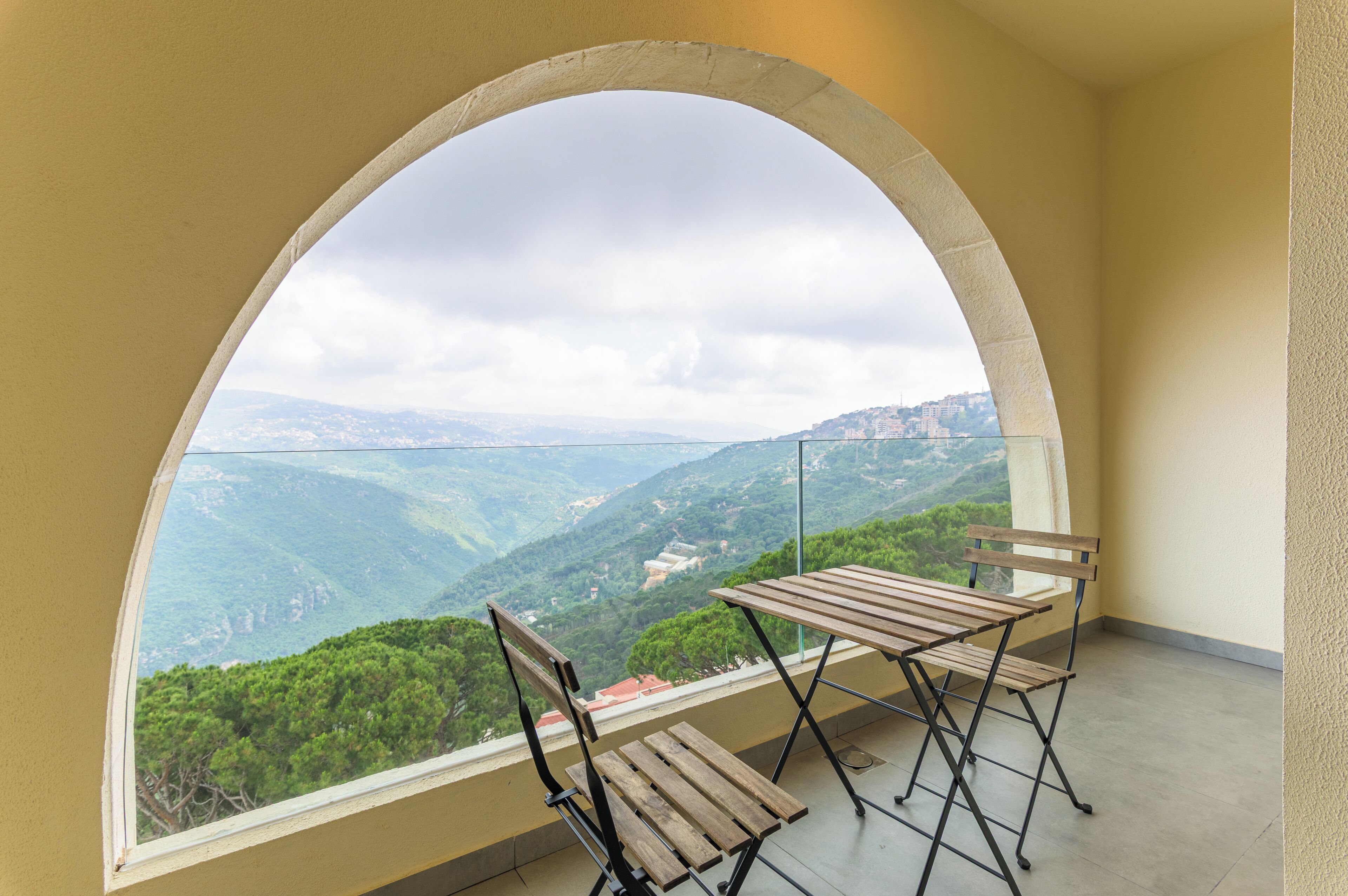 A balcony with a wooden table and chairs, a glass railing, and a view of a mountainous landscape.